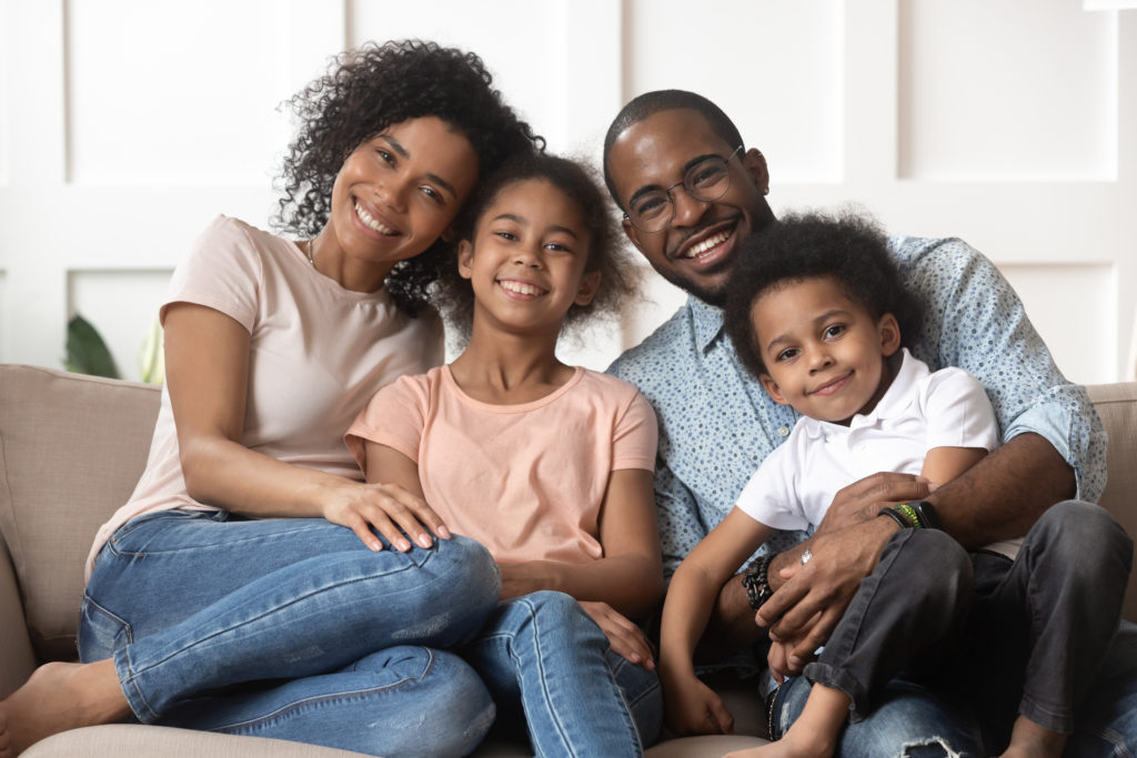 Portrait of happy young african American family with little kids sit relax on couch cuddling, smiling black parents rest on sofa hug preschooler children posing for picture at home together