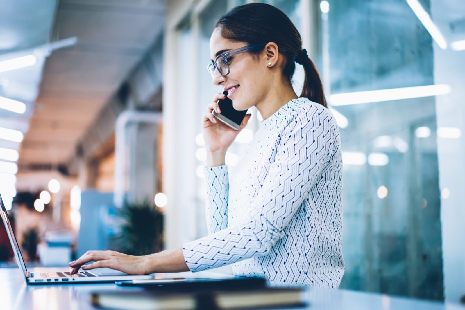 Smiling female receptionist talking on telephone confirmining online booking of client on laptop computer in clinic, positive administrative manager organizing schedule via cellphone conversation